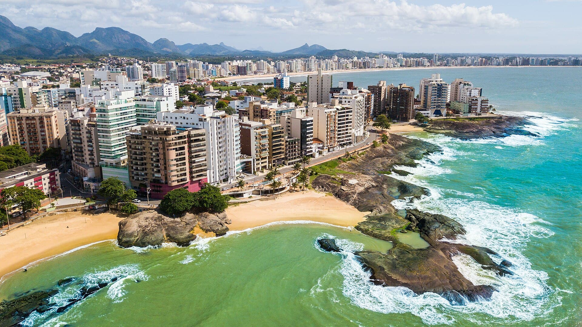 Vista aérea de Guarapari: prédios à beira-mar, uma praia de areia dourada e rochas com ondas